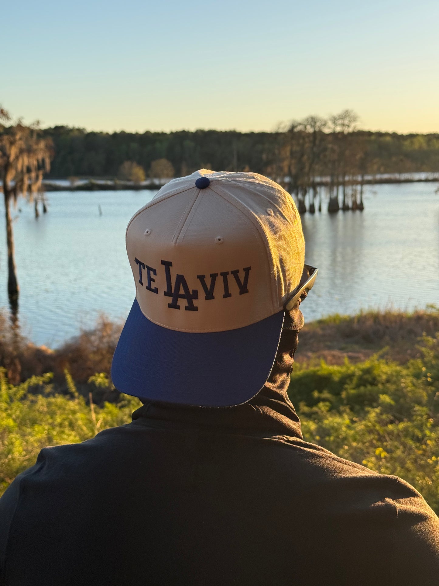 A Chud wearing a cap with 'Tel Aviv' text, standing by a lake at sunset.